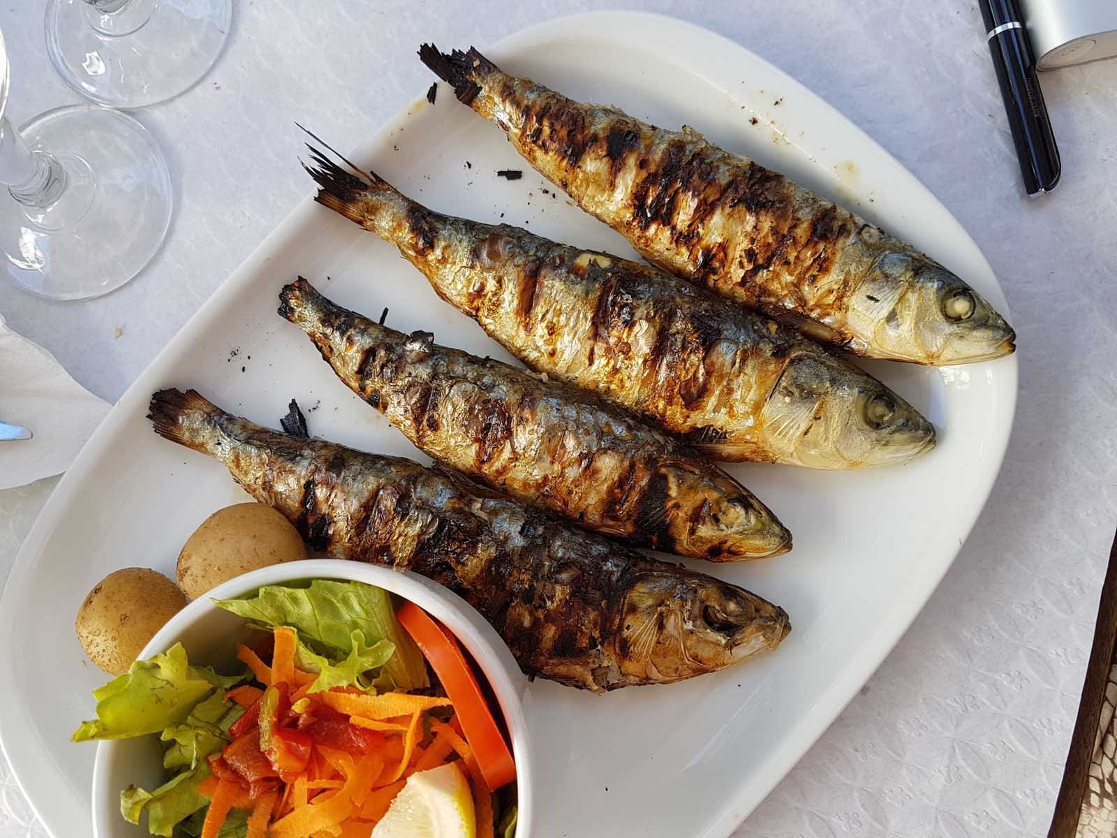 A plate of freshly grilled sardines, a popular food in Portugal, served with a side salad and lemon.